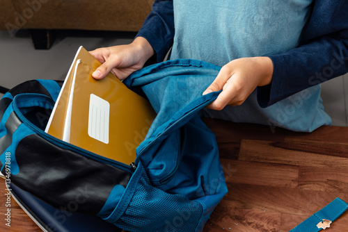 kids placing a yellow notebook inside a blue backpack on a wooden desk, back to school concept