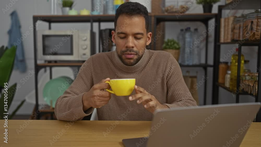 Young man in office drinking coffee from yellow cup in front of laptop in cozy indoor setting with modern kitchen shelves in background, looking focused and relaxed. Young man in office drinking coffee from yellow cup in front of laptop in cozy indoor setting with modern kitchen shelves in background, looking focused and relaxed.