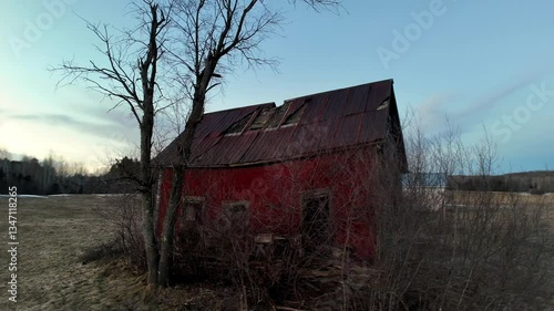 A rustic red barn with a damaged roof stands alone in a field, surrounded by trees and sparse vegetation as evening approaches, casting a serene atmosphere.