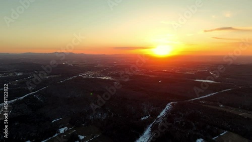 Dramatic sunset paints the sky with warm hues as the sun descends over lush hills and open fields, creating a serene atmosphere in a rural area.