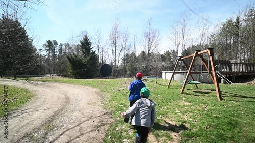 Two boys enjoy their time outdoors in a peaceful rural setting. They are playing and laughing near a parked car, surrounded by trees and a swing set.