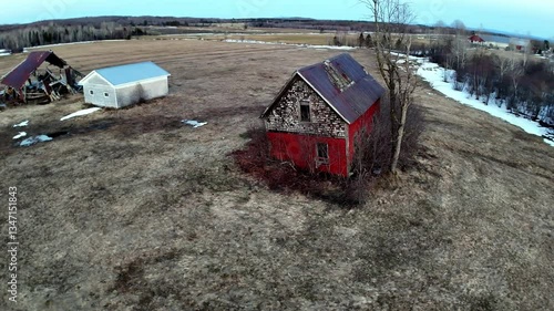 An abandoned red house stands alone in a sprawling rural landscape with patches of snow. Surrounding fields stretch out towards the horizon, creating a serene yet lonely atmosphere.