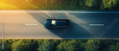 A sleek black car drives along a smooth road surrounded by lush green trees under the golden light of a setting sun from a top-down view

