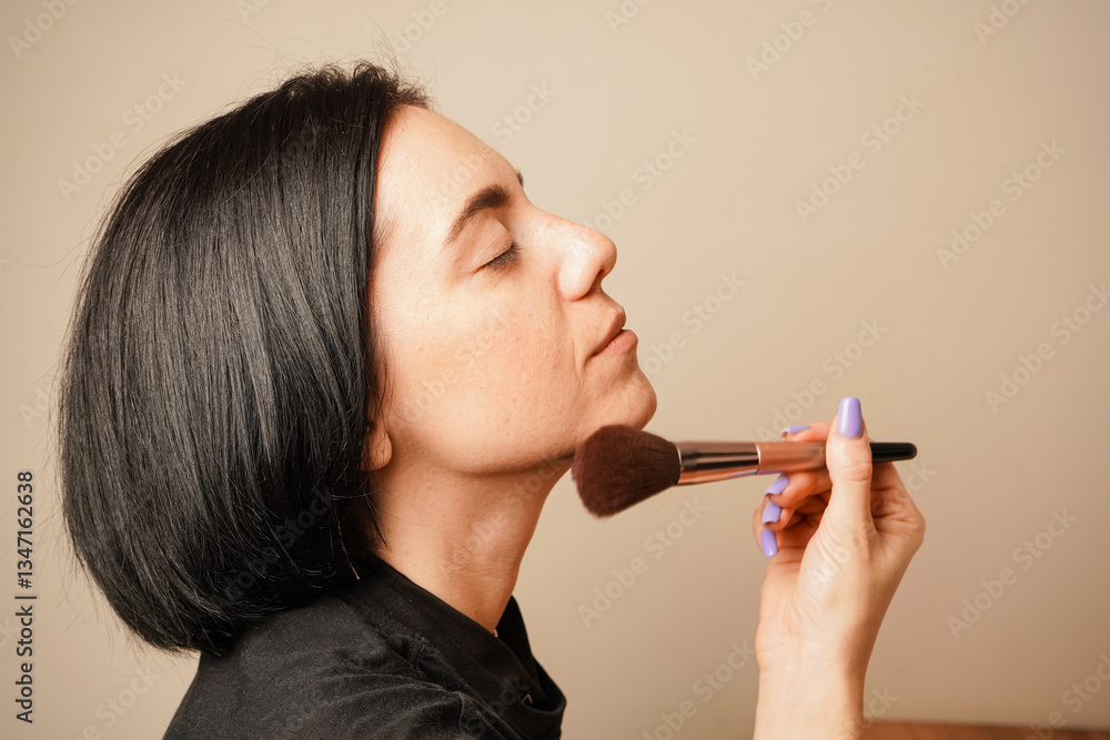 Obraz premium Applying makeup with a soft brush, a woman focuses on perfecting her look. The warm lighting complements her natural skin tone. Her dark hair and serene expression create a calm atmosphere.