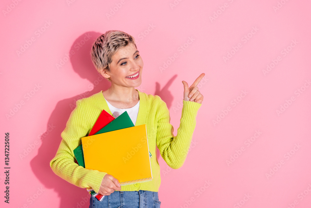 Young woman in trendy casual outfit holding colorful notebooks and pointing with a cheerful expression against a pastel pink background Young woman in trendy casual outfit holding colorful notebooks and pointing with a cheerful expression against a pastel pink background