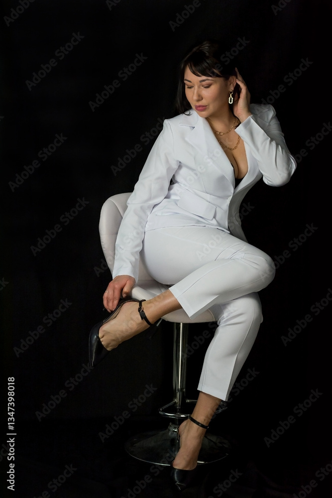 Young pretty woman in a black bra and a business white suit sits on a chair at a black background