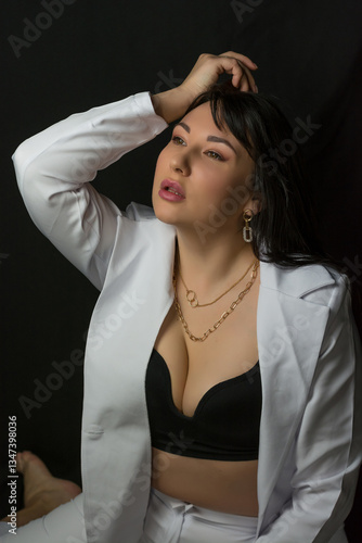 Young pretty brunette woman in a black bra and a business white suit sits at a black background