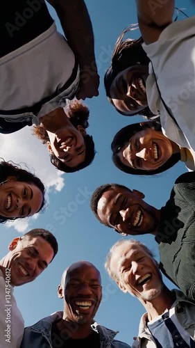 Happy multicultural group of people smiling warmly in a circle, viewed from below against the sky, emphasizing friendship, unity, and happiness.