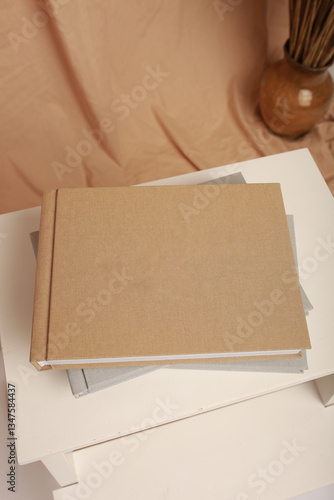 Three elegant photo books with fabric covers are placed on a white chair against a beige fabric backdrop with dried plants. Minimalist design, natural tones, and a cozy atmosphere.