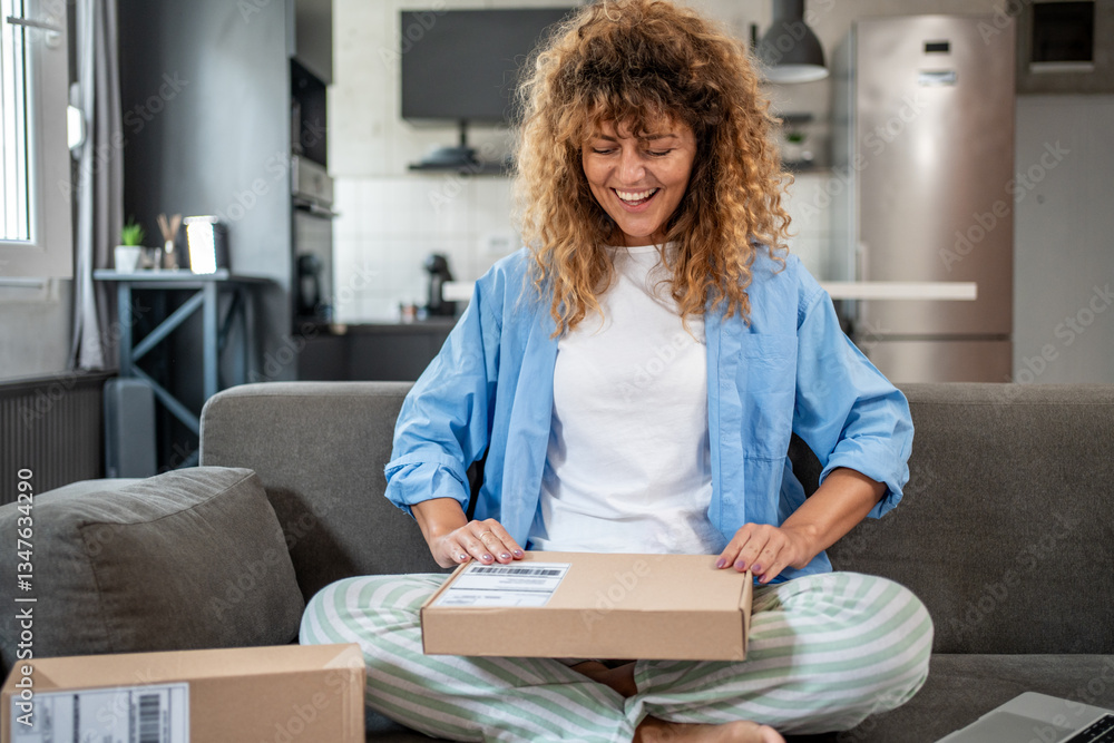 Fototapeta premium Happy woman opening parcel at home on sofa: express delivery and online shopping