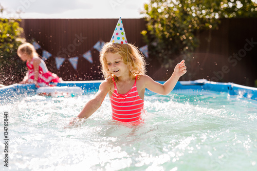 Curly haired blonde girl splashing water in the pool during a party, wearing a paper party hat