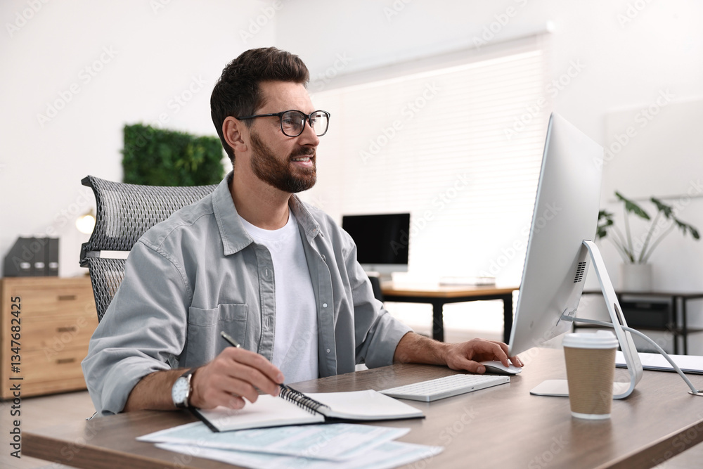 Man taking notes while working on computer at table in office Man taking notes while working on computer at table in office