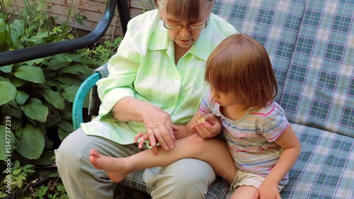 A caring grandmother sits on an outdoor cushioned sofa, gently applying ointment to her young granddaughters leg to soothe a mosquito bite