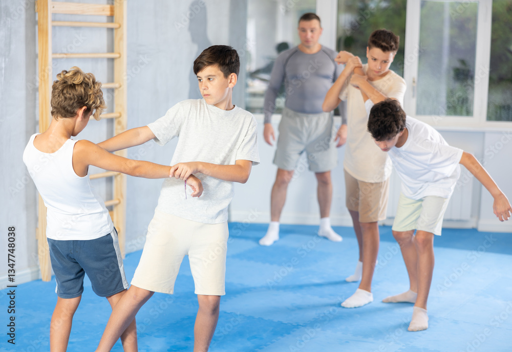 Naklejka premium Focused teenage boys practicing close combat techniques in sparring in training room during self-defence workout under supervision of coach