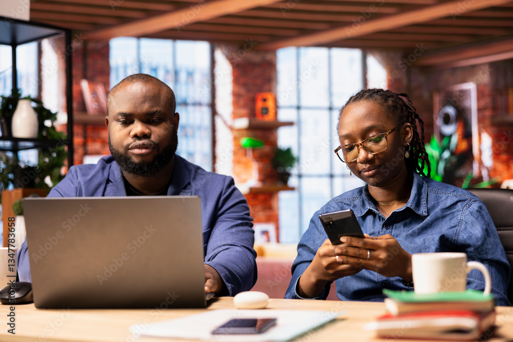 Black couple working remotely on daily freelancing tasks at a desk, young man studying reports on laptop and woman networking with clients on smartphone. Teamwork for efficiency. Black couple working remotely on daily freelancing tasks at a desk, young man studying reports on laptop and woman networking with clients on smartphone. Teamwork for efficiency.