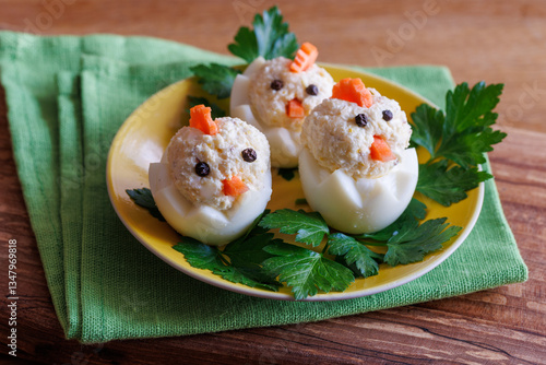 Fun and creative appetizer crafted three egg chick shape. Stuffed boiled eggs with tuna, carrot pieces, and peppercorns on yellow plate with parsley. Easter celebration
