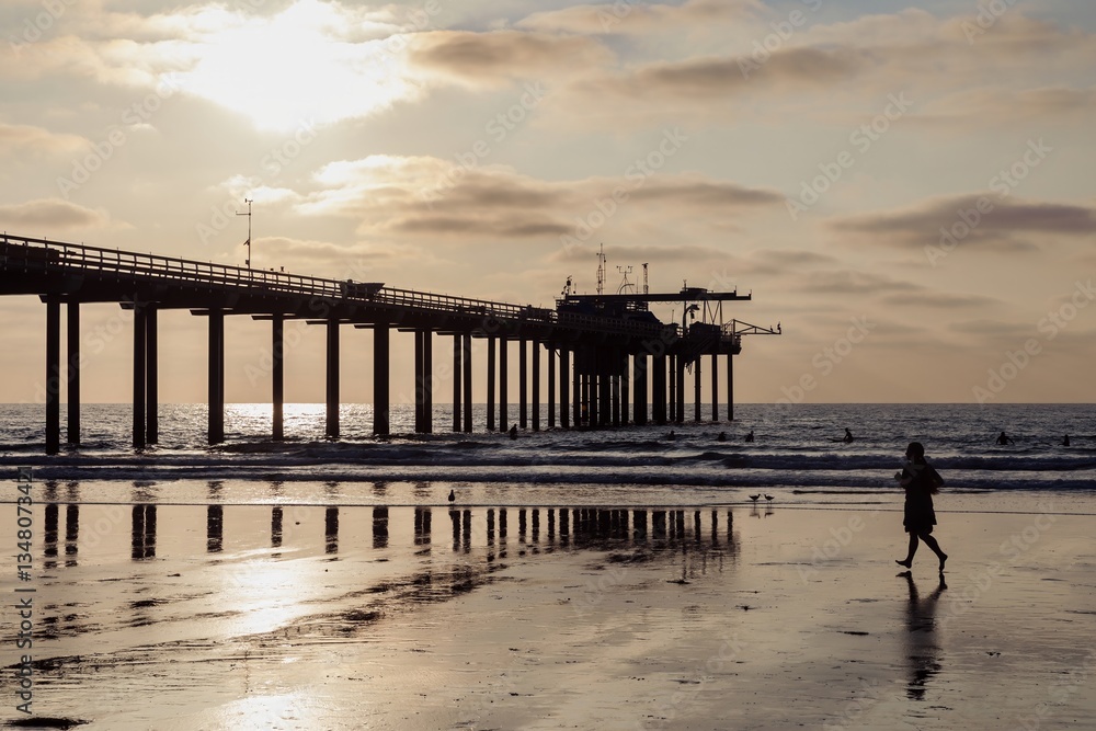 Fototapeta premium A woman jogs along La Jolla beach at sunset in San Diego, California, USA. Surfers are in the water near the Ucsd Scripps Pier, enjoying the waves.