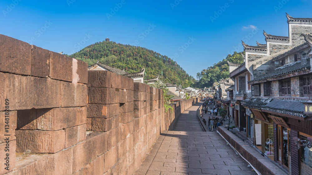 Fototapeta premium The ancient brick fortress wall of Fenghuang Old Town. Traditional Chinese buildings along a paved street. People are walking on the sidewalk. The mountain against the blue sky. China.