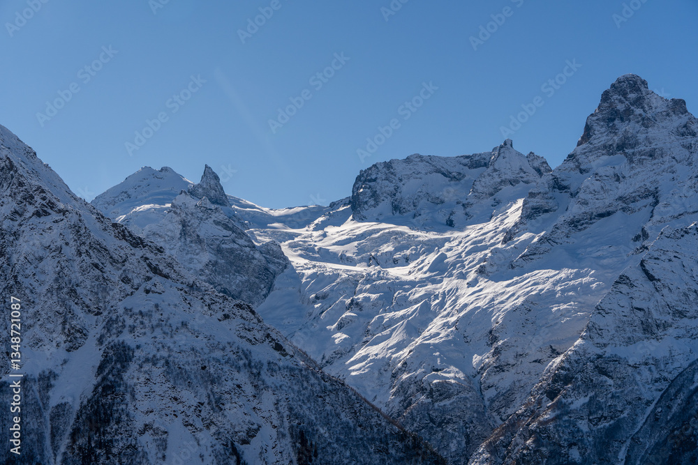 Fototapeta premium Snow-capped peaks with majestic blue slopes rise above jagged slopes of North Caucasus Range. Mountain tops are covered with eternal glaciers. Dombay. Karachay-Cherkessia.