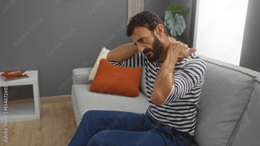Young hispanic man with a beard sits on a couch in a living room massaging his neck while wearing a striped shirt and blue jeans with a relaxed interior setting in the background. Young hispanic man with a beard sits on a couch in a living room massaging his neck while wearing a striped shirt and blue jeans with a relaxed interior setting in the background.