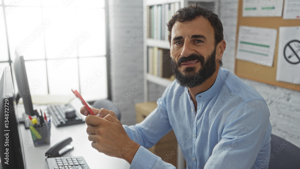 Young, hispanic, man with a beard in an office setting holding a phone, showing a positive expression in a well-lit and organized workplace with documents on a corkboard in the background Young, hispanic, man with a beard in an office setting holding a phone, showing a positive expression in a well-lit and organized workplace with documents on a corkboard in the background