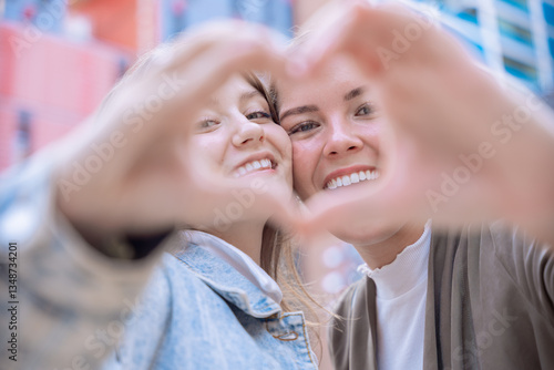 Two happy Caucasian women smiling and making a heart shape with their hands. Symbol of friendship, love, and positivity in a modern urban setting with natural light and joyful expressions.