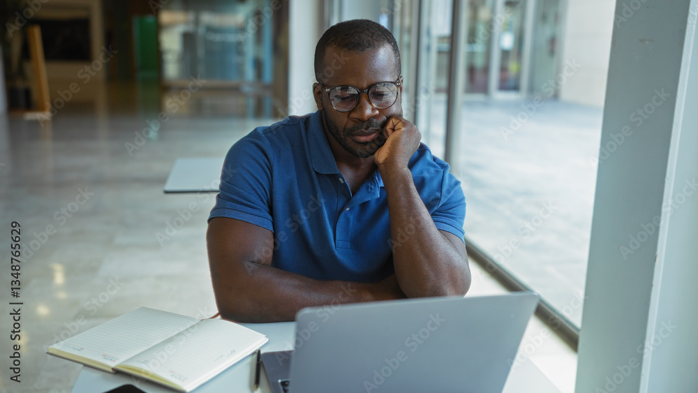 Young man focused on his laptop while working in an office setting with a notebook open on the table Young man focused on his laptop while working in an office setting with a notebook open on the table