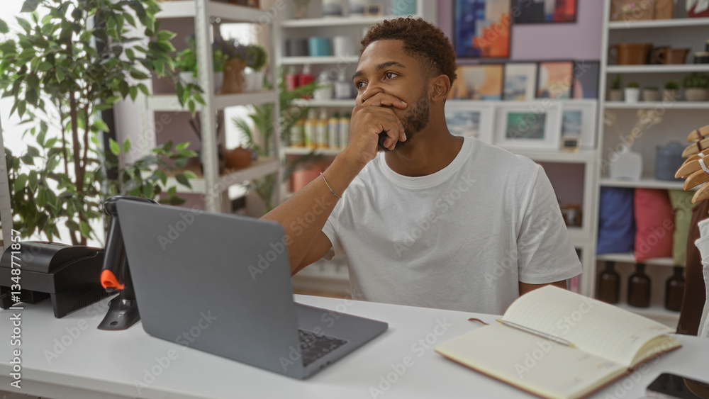 Obraz premium Young man sitting in a home decor store, thoughtfully looking at a laptop while seated at a desk with greenery and decorative items in the background