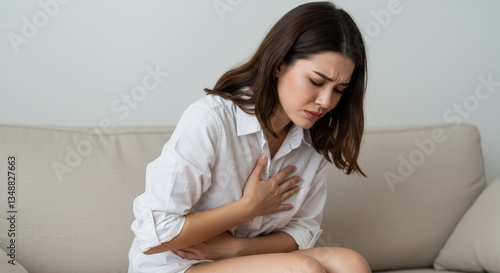 A woman sitting on the couch, clutching her chest in pain while wearing a white shirt.