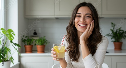 A happy woman holding a refreshing lemon drink in a glass, smiling and resting her hand on her cheek in a cozy kitchen with plants and natural light.
