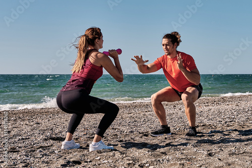 Personal trainer guiding woman doing squats with dumbbells on beach