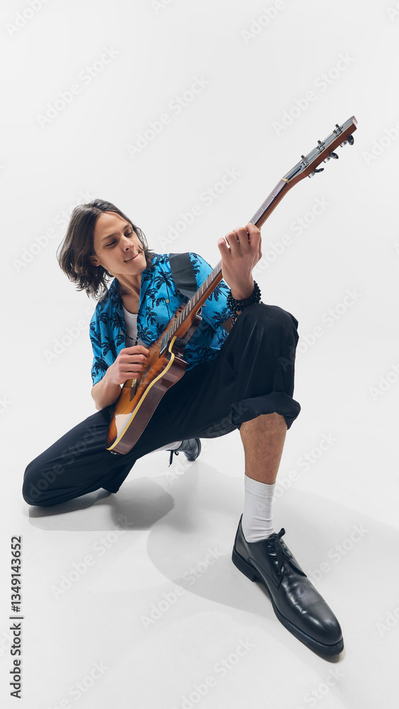 Fototapeta premium Young energetic man in tropical shirt, sitting and passionately playing electric guitar against white background. Concept of music, performance, festival, talent, passion, artistic movement.