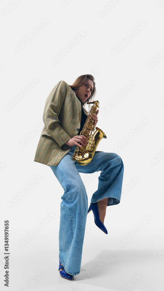 Naklejka premium Stylish young woman in vintage blazer and wide-leg jeans, lifting leg while passionately playing golden saxophone on white background. Concept of music, jazz, festival, stage movement, freedom.