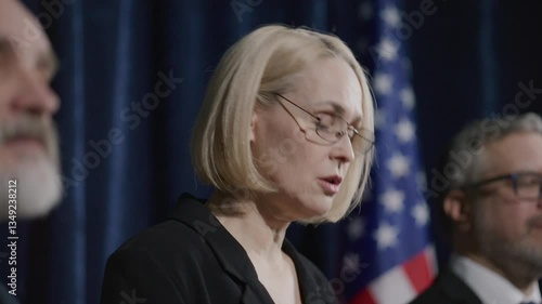 Female senator standing on stage with two male colleagues, delivering speech to audience during political press conference with US flag in background