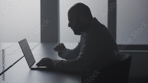 Man fertigt Bericht im Büro an Schreibtisch auf Laptop an, sitzt im Schatten mit Gegenlicht der Fenster
