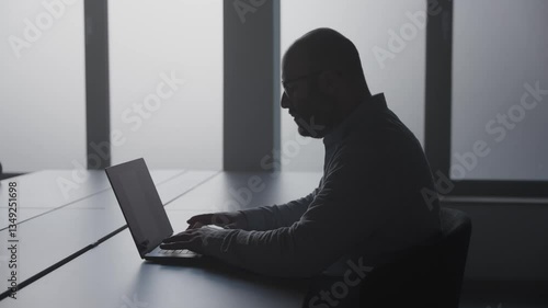 Man fertigt Bericht im Büro an Schreibtisch auf Laptop an, sitzt im Schatten mit Gegenlicht der Fenster
