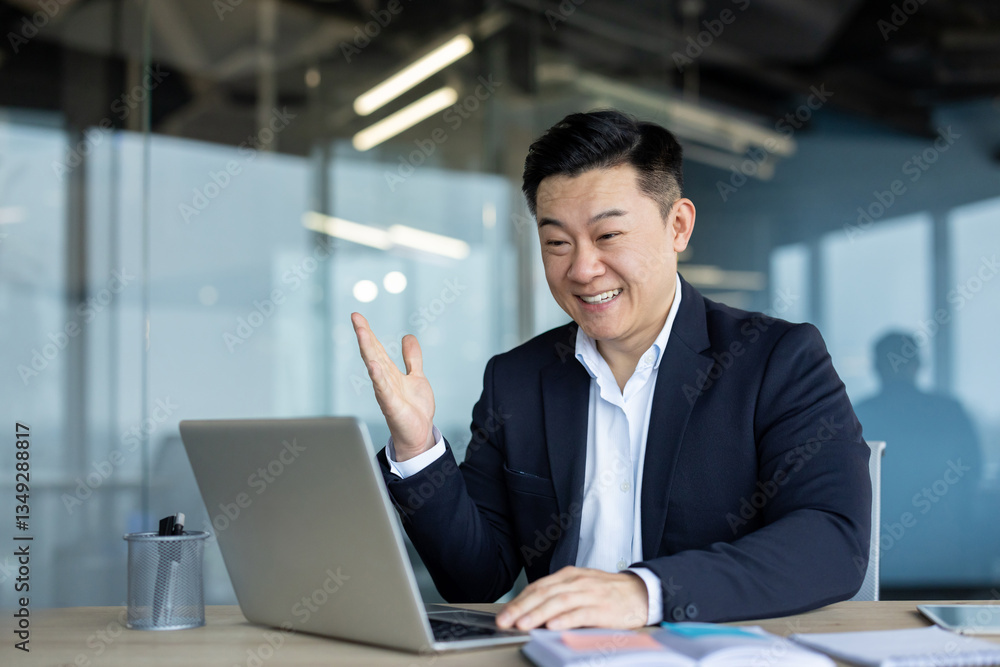 Happy young Asian man in a business suit sitting at a desk in an office, looking at a laptop screen and rejoicing in success and news Happy young Asian man in a business suit sitting at a desk in an office, looking at a laptop screen and rejoicing in success and news
