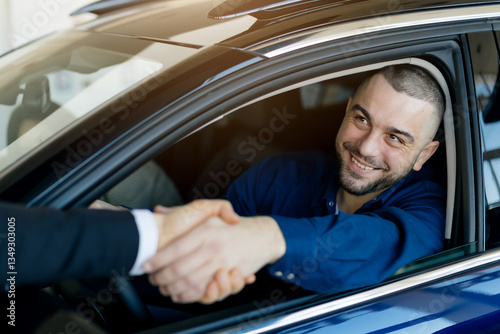 A happy male customer sits inside his newly purchased car at a modern dealership showroom, shaking hands with the salesman through the open window, having positive car buying experience