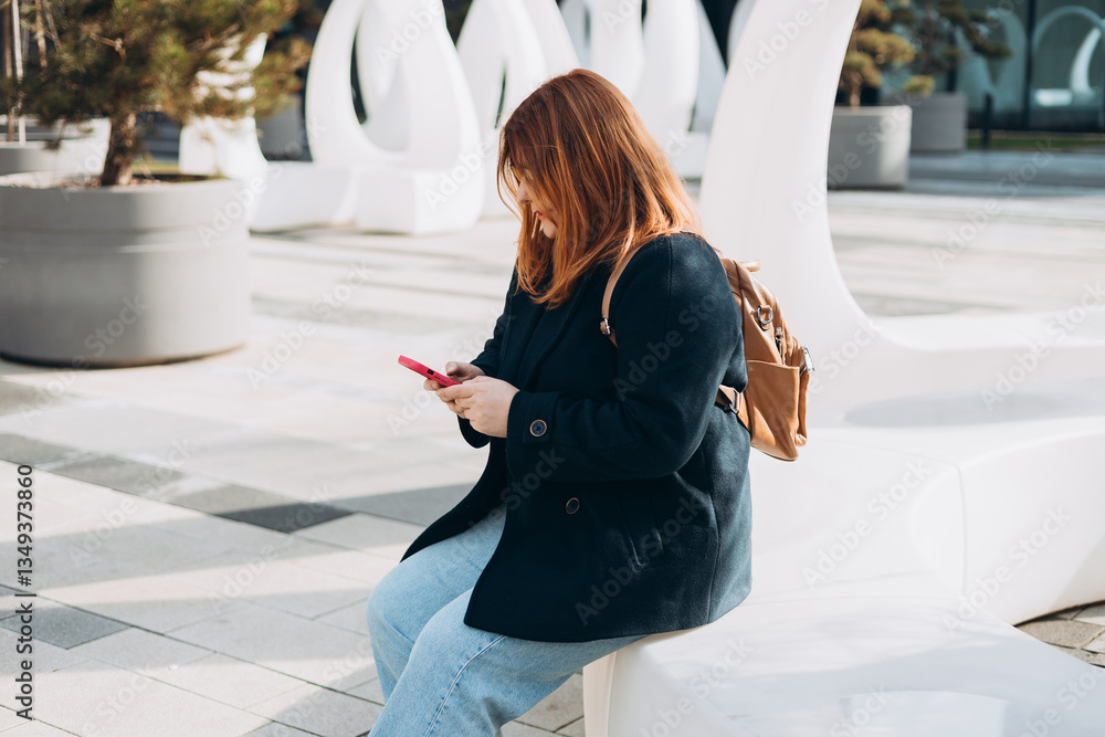 Happy woman chatting on phone and relaxing outdoors. Urban, people concept. Smiling 30s redhead woman text messaging on smart phone at sunny day on the city street, spring time