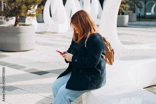 Obraz na płótnie Happy woman chatting on phone and relaxing outdoors. Urban, people concept. Smiling 30s redhead woman text messaging on smart phone at sunny day on the city street, spring time w salonie