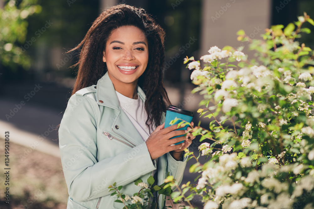 Obraz premium Charming young woman enjoying a sunny day outdoors on a city walk, holding a coffee cup and smiling near blooming flowers