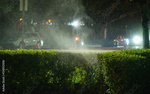 Sprinkler system operates at night creating mist near a hedge with shining car headlights in the background