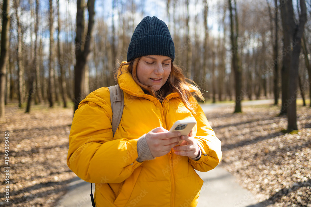 A happy woman in a yellow jacket and black beanie walking in the park, holding her phone. The bright natural setting, with trees and a peaceful path, enhances the cheerful mood of the moment.