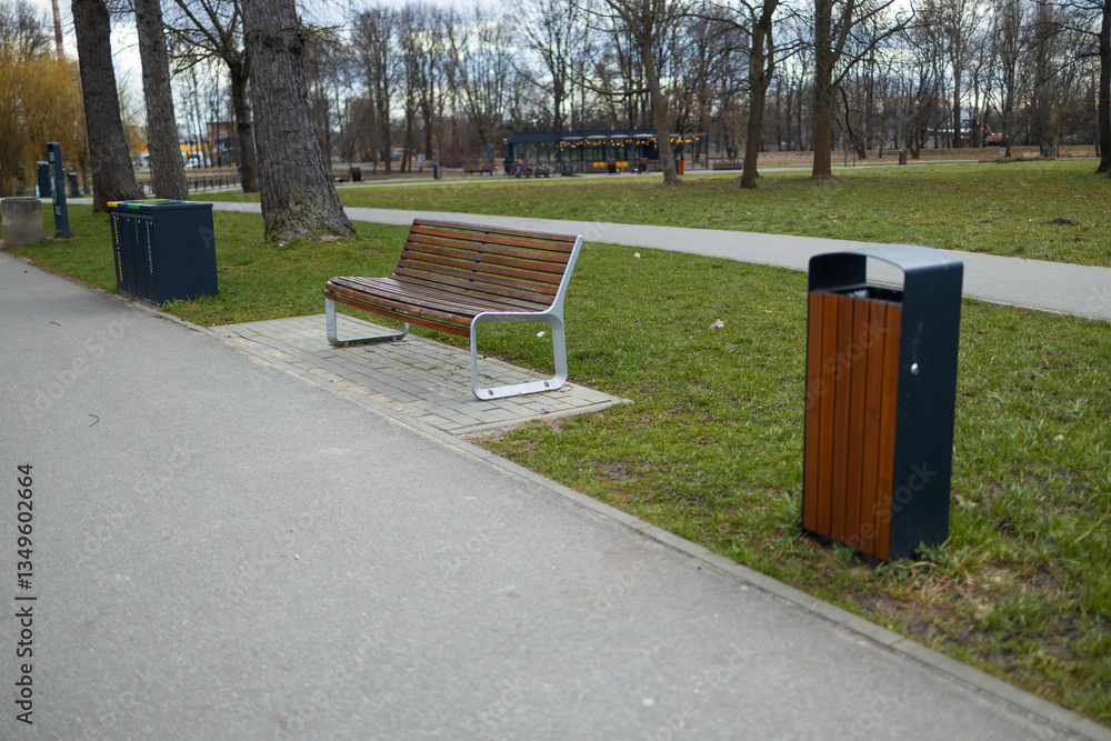 A well-maintained public park scene featuring a wooden bench, trash bins, and a green park setting. The path runs through the park with trees in the background, offering a peaceful atmosphere