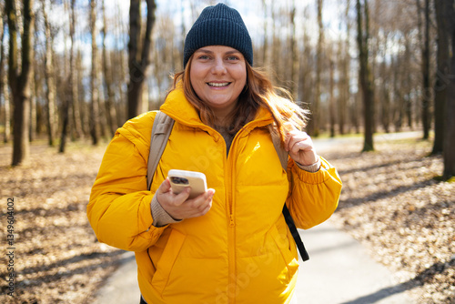 A happy woman in a yellow jacket and black beanie walking in the park, holding her phone. The bright natural setting, with trees and a peaceful path, enhances the cheerful mood of the moment.