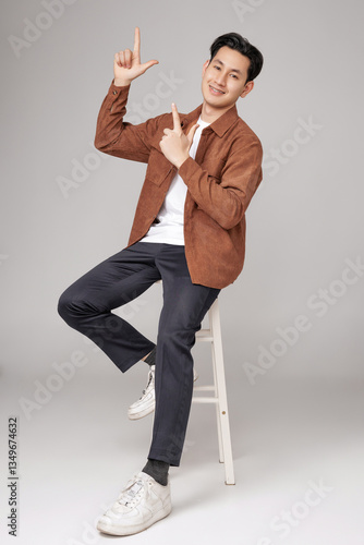 Young Asian man dressed casually in a brown shirt and white sneakers pointing finger to copy space on isolated background. His cheerful relaxation and approachability.