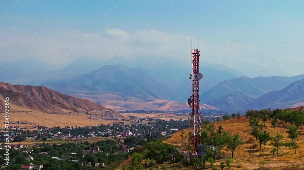 Drone footage showing a mountain village near with a telecom tower on a dry hill. The drone slowly passing by a tower with distant mountains in the background at late summer day.