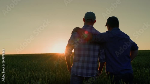 Father and son in a field at sunset. The son is slightly in front, wearing a brightly colored plaid shirt and baseball cap. His arm is around the shoulders of the man behind him.