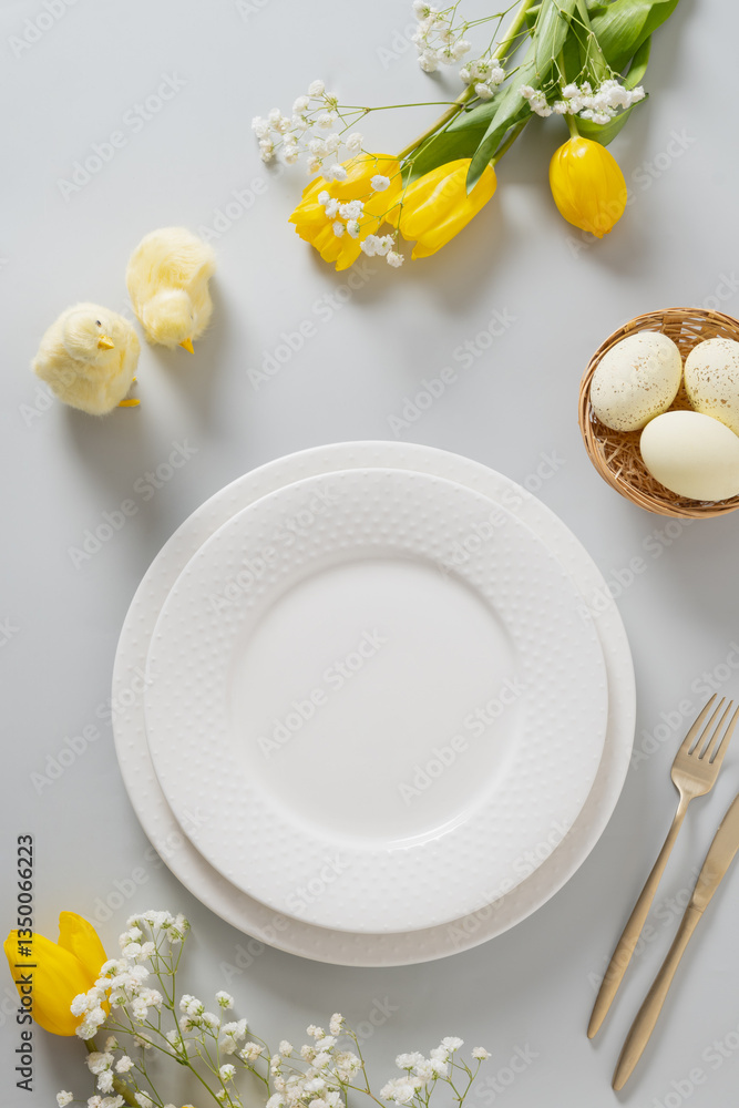 Easter table setting with spring, yellow tulips, white plates, bunny, eggs on white background. Happy Easter celebration. View from above. Vertical format.