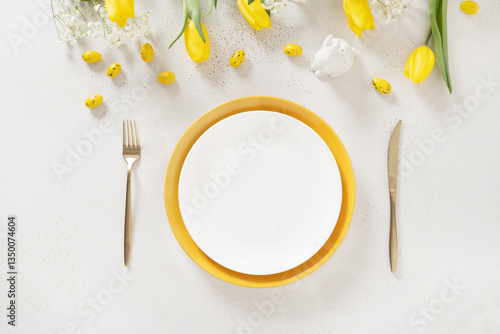 Easter table setting with spring yellow tulips, white plates, bunny, eggs on white background. Happy Easter celebration. Copy space. View from above.